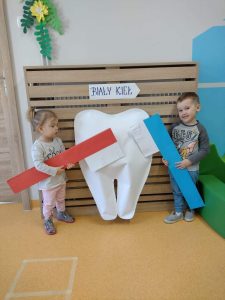 Dzieci stoją koło dużego białego zęba w rączkach trzymają duże papierowe szczoteczki w kolorze niebieskim i czerwonym.
