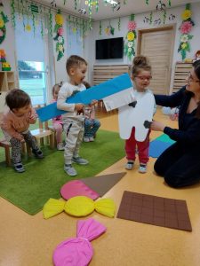 Dziewczynka stoi przebrana za białego chorego ząbka przed nią leżą duże papierowe cukierki, lody i czekolada. Obok na krzesełkach siedzą dzieci i patrzą z wielkim zainteresowaniem. Chłopczyk trzyma w dłoniach dużą niebieską papierową szczoteczkę i czyści chorego ząbka w którego jest przebrana koleżanka. W tle widać sale zabaw.