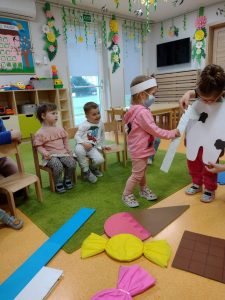 Dziewczynka stoi przebrana za białego chorego ząbka przed nią leżą duże papierowe cukierki, lody i czekolada. Obok na krzesełkach siedzą dzieci i patrzą z wielkim zainteresowaniem. Dziewczynka przebrana za dentystkę leczy chorego ząbka papierowym wiertłem. W tle widać sale zabaw.