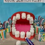 Na środku stolika stoi makieta przedstawiająca jamę ustną, koło niej stoją kolorowe papierowe szczoteczki i kubeczki zrobione przez dzieci. U góry znajduje się napis " Kącik Dentystyczny".