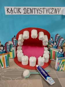 Na środku stolika stoi makieta przedstawiająca jamę ustną, koło niej stoją kolorowe papierowe szczoteczki i kubeczki zrobione przez dzieci. U góry znajduje się napis " Kącik Dentystyczny".