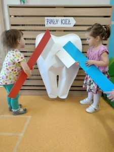 Dzieci stoją i trzymają w dłoniach duże papierowe szczoteczki w kolorze niebieskim i czerwonym, naśladując nimi mycie ząbków na dużym białym zębie.