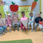 Dzieci siedzą w rzędzie na krzesełkach trzymając w dłoniach duże papierowe owoce, podnosząc je do góry. W tle widać żółty plakat z napisem " Uśmiech od ucha do ucha"