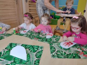 Dzieci siedzą przy stoliku, na zielonych podkładkach wyklejają białe serduszko czerwonymi kółeczkami tworząc z nich flagę polski. W tle widać opiekunkę nachyloną nad chłopcem w krzesełku, który wykonuje prace plastyczną.
