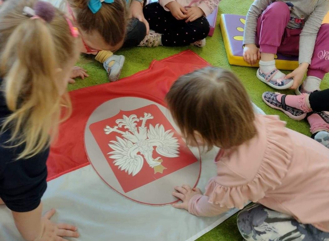 Dzieci siedzą po turecku na zielonym dywanie między nimi jest rozłożona flaga polski i godło. Dzieci patrzą i oglądają z wielkim zaciekawieniem.