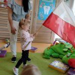 Dziewczynka stoi na zielonym dywanie i trzyma w dłoniach dużą flagę polski, wymachując nią. Opiekunka stoi za dziewczynką.