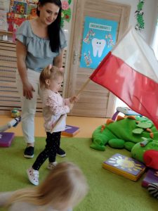 Dziewczynka stoi na zielonym dywanie i trzyma w dłoniach dużą flagę polski, wymachując nią. Opiekunka stoi za dziewczynką.