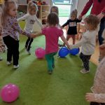 Dzieci trzymają się za ręce i tańczą w kółeczku. Dookoła leża kolorowe baloniki.