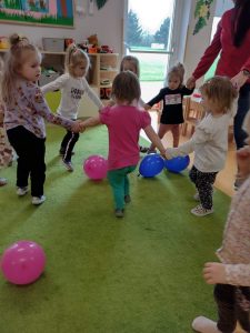 Dzieci trzymają się za ręce i tańczą w kółeczku. Dookoła leża kolorowe baloniki.