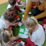 Dzieci siedzą na dywanie ubrane na biało - czerwono i wraz z opiekunką przyklejają białe kwadraty na zielonym kartonie.