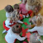 Dzieci siedzą na dywanie ubrane na biało - czerwono i wraz z opiekunką przyklejają czerwone kwadraty na zielonym kartonie.