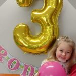 Opiekunka trzyma dziewczynkę na rękach., która trzyma w dłoniach różowy balonik. W tle widać napis " Urodziny" i duży balon w kształcie cyfry trzy w złotym kolorze.