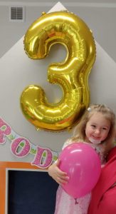 Opiekunka trzyma dziewczynkę na rękach., która trzyma w dłoniach różowy balonik. W tle widać napis " Urodziny" i duży balon w kształcie cyfry trzy w złotym kolorze.