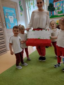 Opiekunka ubrana na biało - czerwono trzyma w dłoniach dużą papierową flagę zrobioną przez dzieci. Obok niej stoją dzieci ubrane na biało- czerwono.