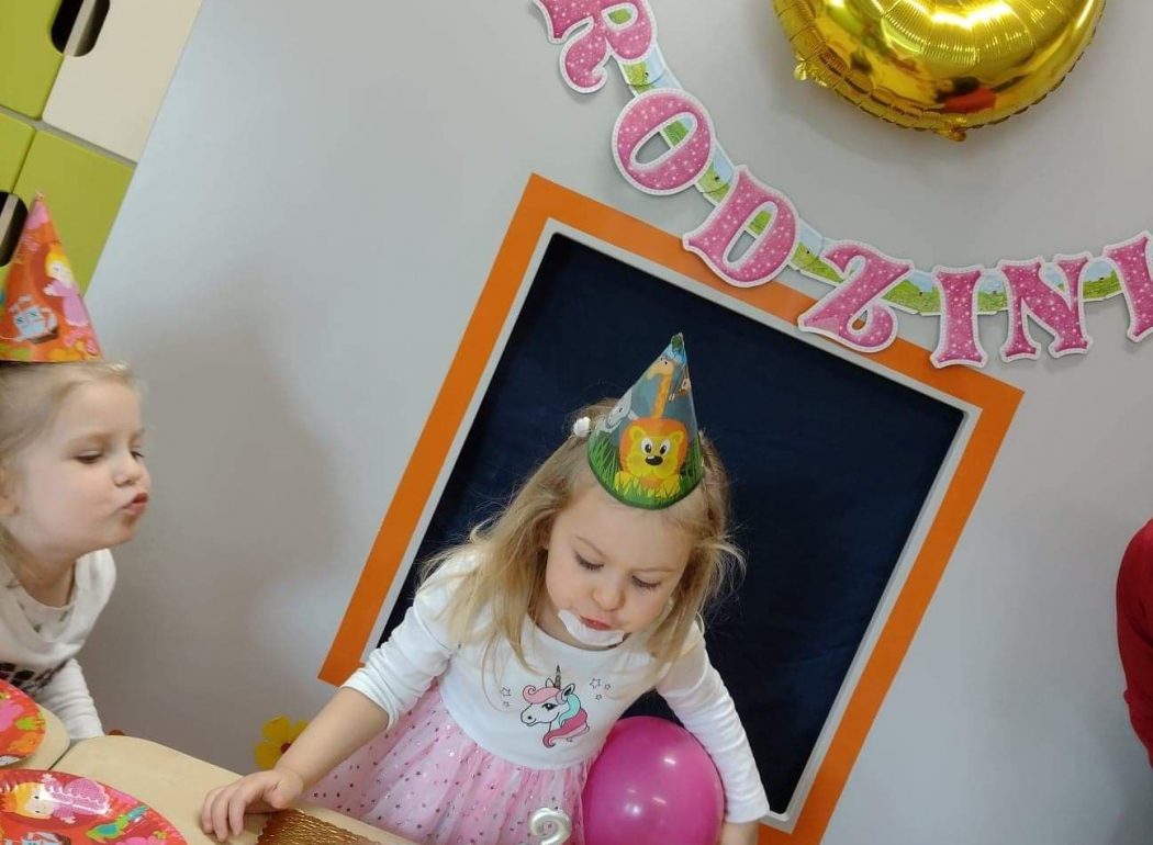 Dziewczynka stoi przed stołem i dmucha na świeczkę w kształcie cyfry trzy wbitej do urodzinowego torciku z babeczek. W tle widać napis "Urodziny" i duży balon w kształcie cyfry trzy w złotym kolorze.