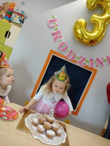 Dziewczynka stoi przed stołem i dmucha na świeczkę w kształcie cyfry trzy wbitej do urodzinowego torciku z babeczek. W tle widać napis "Urodziny" i duży balon w kształcie cyfry trzy w złotym kolorze.