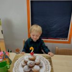 Chłopiec siedzi przy stoliku ubrany w ciemno- granatową koszulę w kratkę. Na stoliku przed chłopcem stoi torcik zrobiony z muffinek z urodzinową świeczką w kształcie cyfry 2. W tle widać urodzinowy napis.