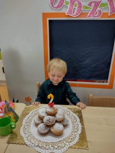 Chłopiec siedzi przy stoliku ubrany w ciemno- granatową koszulę w kratkę. Na stoliku przed chłopcem stoi torcik zrobiony z muffinek z urodzinową świeczką w kształcie cyfry 2. W tle widać urodzinowy napis.