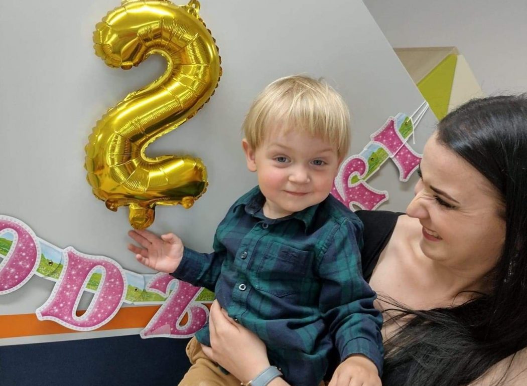 Opiekunka trzyma Solenizanta na rękach. Chłopiec uśmiecha się i trzyma rączką złoty balonik w kształcie cyfry 2. W tle widać urodzinowy napis.
