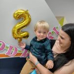 Opiekunka trzyma Solenizanta na rękach. Chłopiec uśmiecha się i trzyma rączką złoty balonik w kształcie cyfry 2. W tle widać urodzinowy napis.