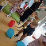 Dzieci wraz z opiekunką tańczą z kolorowymi balonikami. W tle widać sale zabaw.