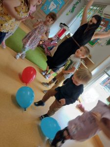 Dzieci wraz z opiekunką tańczą z kolorowymi balonikami. W tle widać sale zabaw.