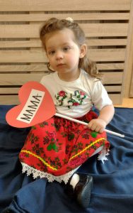 Dziewczynka siedzi po turecku w białej bluzeczce i krakowskiej spódniczce. W rączce trzyma czerwone serduszko z napisem "MAMA".