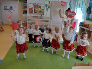 Dzieci tańczą do piosenki, w rączce trzymają na patyczku czerwone serduszko z napisem "MAMA". Dziewczynki ubrane są w białe koszulki i krakowskie spódniczki a chłopcy w strój galowy. W tyle widać sale zabaw.