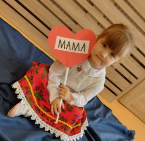 Dziewczynka siedzi po turecku w białej bluzeczce i krakowskiej spódniczce. W rączce trzyma czerwone serduszko z napisem "MAMA".