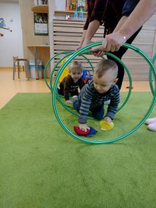 Opiekunki trzymają pionowo hula-hop na zielonym dywanie, tworząc z nich tunel. Dzieci przechodzą pojedynczo przez tunel zrobiony z hula-hop.