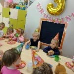 Dzieci ubrane w urodzinowe czapeczki siedzą przy stoliku i częstują się muffinkami. W tle widać urodzinową dekoracje.
