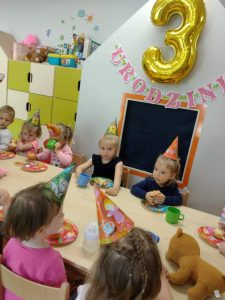 Dzieci ubrane w urodzinowe czapeczki siedzą przy stoliku i częstują się muffinkami. W tle widać urodzinową dekoracje.