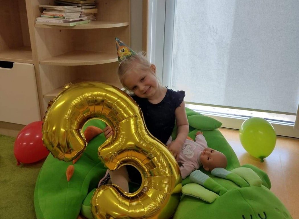 Dziewczynka siedzi na dużym zielonym smoku, w rączce trzyma duży złoty balon w kształcie cyfry trzy.