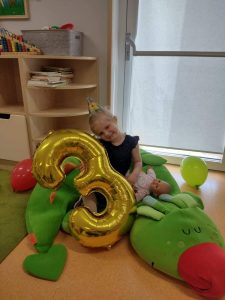 Dziewczynka siedzi na dużym zielonym smoku, w rączce trzyma duży złoty balon w kształcie cyfry trzy.