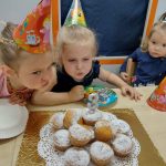 Dziewczynki ubrane w czapeczki urodzinowe siedzą przy stole i dmuchają świeczkę na urodzinowym torcie z muffinek.