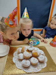 Dziewczynki ubrane w czapeczki urodzinowe siedzą przy stole i dmuchają świeczkę na urodzinowym torcie z muffinek.