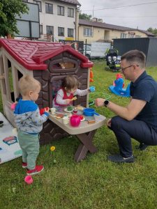 Pan Wójt kuca przy zabawkowym domku ogrodowym i bawi się z dziewczynką i chłopcem w gotowanie.