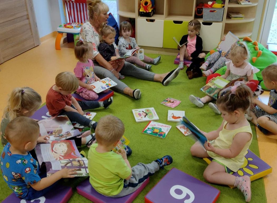 Dzieci siedzą wraz z ciocią na zielonym dywanie. Na środku rozłożone są książeczki. Dzieci oglądają książeczki z wielkim zainteresowaniem.