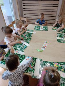 Dzieci siedzą przy stoliku i na zielonych podkładkach wyklejają kolorowymi kwiatuszkami zakładki do książeczek.