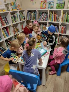 Dzieci siedzą w bibliotece na niebieskich krzesełkach przy stoliku i oglądają książeczki. W tle widać regał z książeczkami.