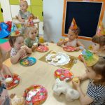Dzieci siedzą przy stoliku ubrane w urodzinowe czapki i częstują się muffinkami.