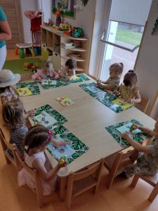 Dzieci siedzą przy stoliku i przyklejają na kartce elementy związane z plażą.