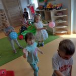 Dzieci tańczą z kolorowymi balonikami na zielonym dywanie. W tle widać sale zabaw.