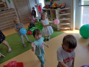 Dzieci tańczą z kolorowymi balonikami na zielonym dywanie. W tle widać sale zabaw.