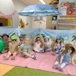 Dzieci ubrane w letnie kapelusze i okulary siedzą i pozują do zdjęcia. W tle widać letnią scenerię, duży plażowy parasol i parawan.