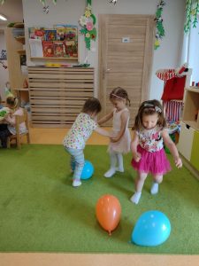 Dziewczynki tańczą z kolorowymi balonikami na zielonym dywanie. W tle widać sale zabaw.