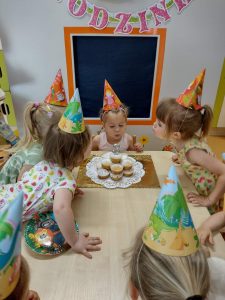 Dzieci ubrane w urodzinowe czapeczki siedzą przy stoliku obok solenizantki i zdmuchują świeczkę na muffinkowym torcie.