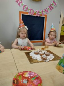 Dziewczynki siedzą przy stoliku urodzinowym. Na środku stolika leży torcik zrobiony z muffinek. W tle widać dekoracje urodzinową.