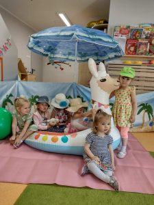 Dzieci ubrane w letnie kapelusze i okulary pozują do zdjęcia siedzą na dużej dmuchanej lamie. W tle widać letnią scenerię, duży plażowy parasol i parawan.