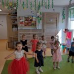 Dzieci stoją na zielonym dywanie ubrane odświętnie i tańczą do piosenki pt." Tato, Tato" . W tle widać sale zabaw.